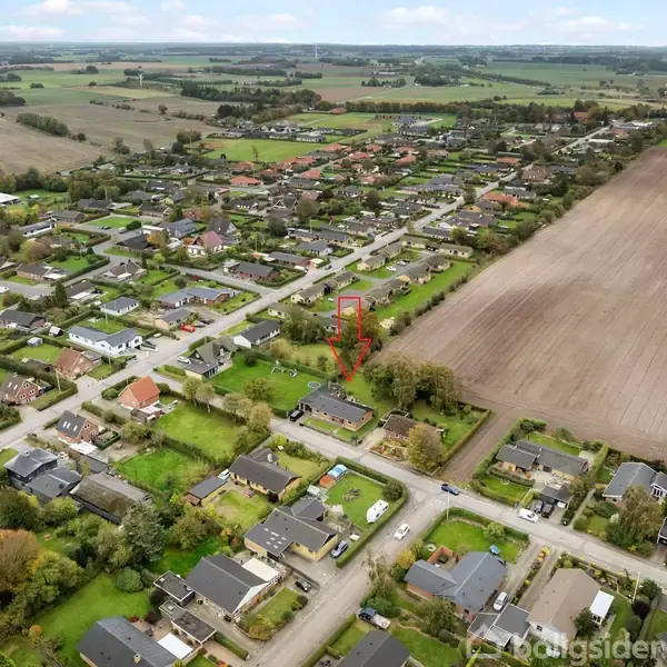 Et boligområde med spredte huse tæt på marker. En rød pil peger mod et specifikt hus. Området er omgivet af landligt landskab og åbne marker.