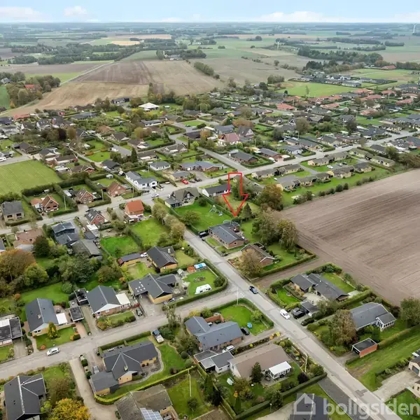 Landsby med spredte huse og veje set fra oven, omgivet af marker og grønne områder. En rød pil markerer et specifikt sted blandt husene. Skyer pryder den blå himmel.