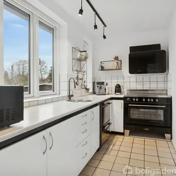 Et moderne køkken med hvide skabe og sorte bordplader har en opvaskemaskine og mikroovn til venstre. I baggrunden står en komfur og en ovn ved siden af flisbelagte vægge.