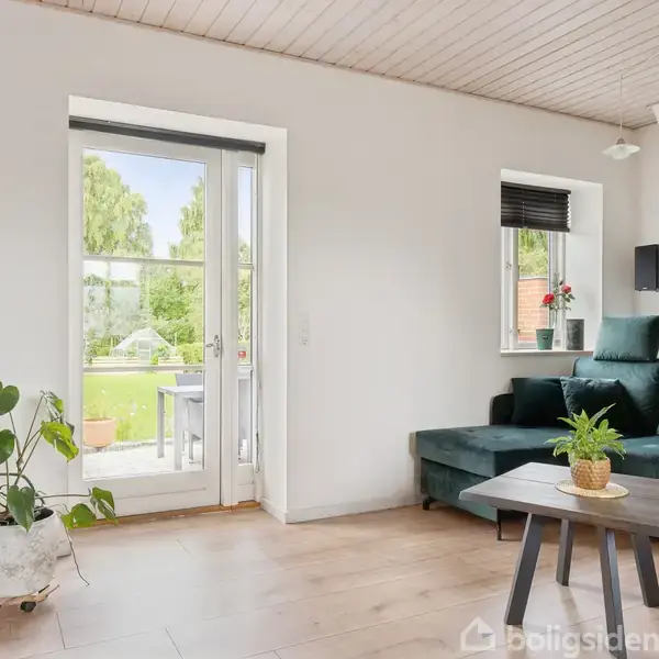En grøn sofa står i hjørnet af en lys stue ved siden af en glasdør, der åbner ud til en terrasse. Et lille bord med en plante står foran sofaen.