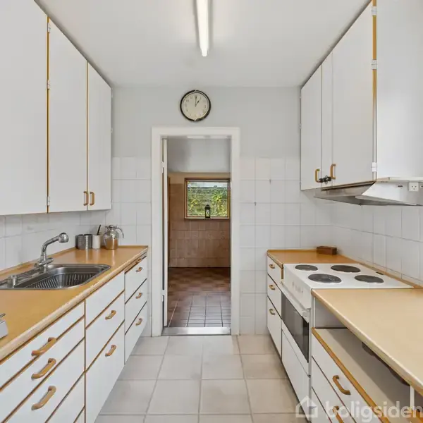 Et køkken med hvide skabe og brune bordplader, en indbygget kogeplade og en vask. Rummet er smalt med flisegulv. Gennem en dør ses et tilstødende rum med et vindue.