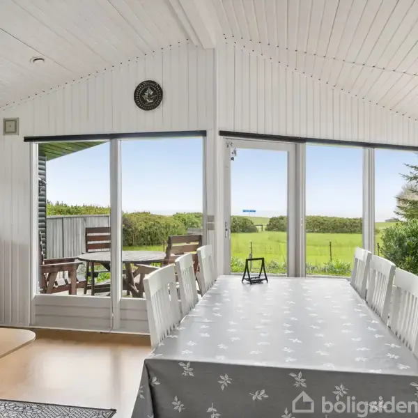Et spisebord med dug står i en lys stue med hvide trævægge. Udenfor ses en terrasse med stole og grønne marker, der strækker sig mod horisonten.