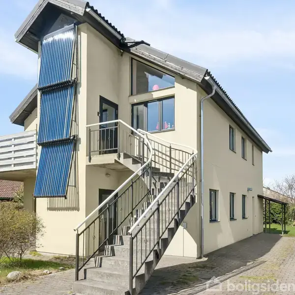 Haus med synlige solpaneler på en skrå vægdel og udendørs trappe, der leder til en balkon. Omkringliggende haver og andre huse i nabolaget ses under en klar blå himmel.