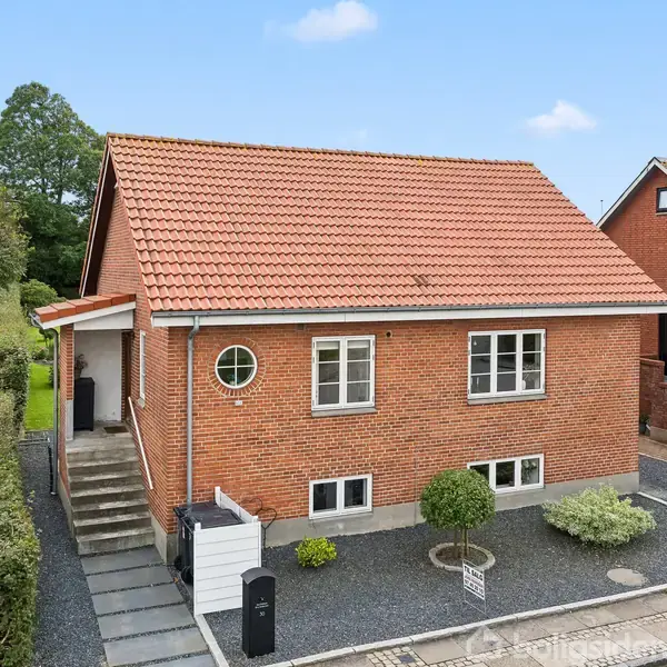 Et rødstenshus med rødt tag står i et beboelsesområde. En have med grus og planter omringer huset, og en gangsti fører op til hoveddøren.