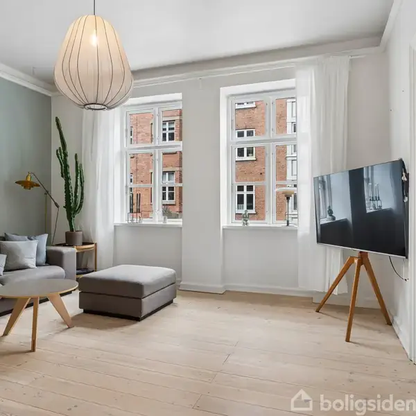 Grå hjørnesofa med puder står i en lys stue med trægulve. En stor lampe hænger ned fra loftet. Et fladskærms-TV på stativ står ved siden af et vindue.
