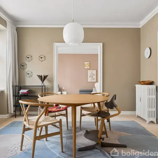 Et rundt træbord omgivet af stole står i en lys stue. En hvid lampe hænger over bordet, omgivet af beige vægge, et vindue, og åbne døre til et tilstødende rum.