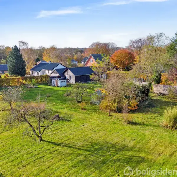 Grøn have med spredte træer, der omkranser et gråt hus. Omgivende område inkluderer flere huse og træer i efterårsfarver under en blå himmel.