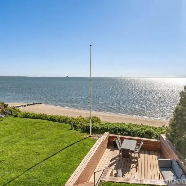 Terrasse med møbler vender ud mod sandstrand og hav under en klar blå himmel. Græsplæne og buske omgiver området, skaber en fredelig kystatmosfære.