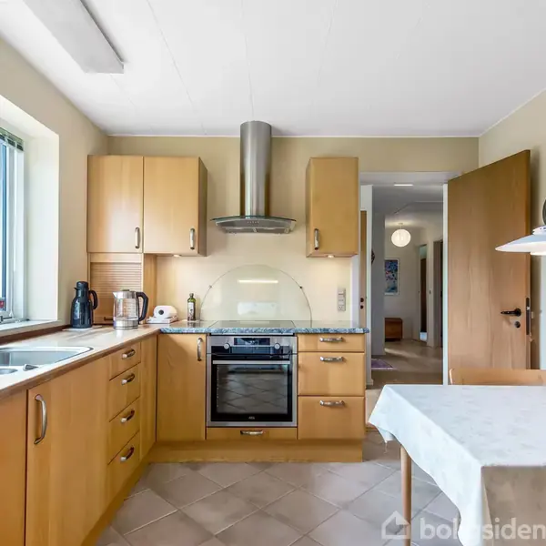 Køkken med træskabe omkranser moderne komfur og emhætte, mens et spisebord med hvid dug står til højre. Et stort vindue giver udsigt til grøn bevoksning udenfor.