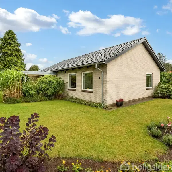 Et enkelt etplanshus med hvid murstensfacade og gråt tegltag står i en velplejet have med grønt græs og forskellige buske.