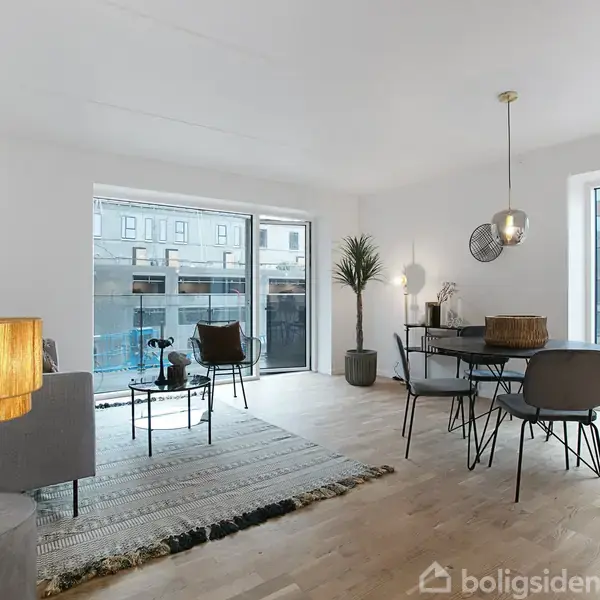 Stue med lyse trægulve, hvor en grå sofa og to stole står omkring et tæppe. En gulvlampe lyser rummet, der indeholder et rundt spisebord med stole. Store vinduer viser moderne bybyggeri udenfor.