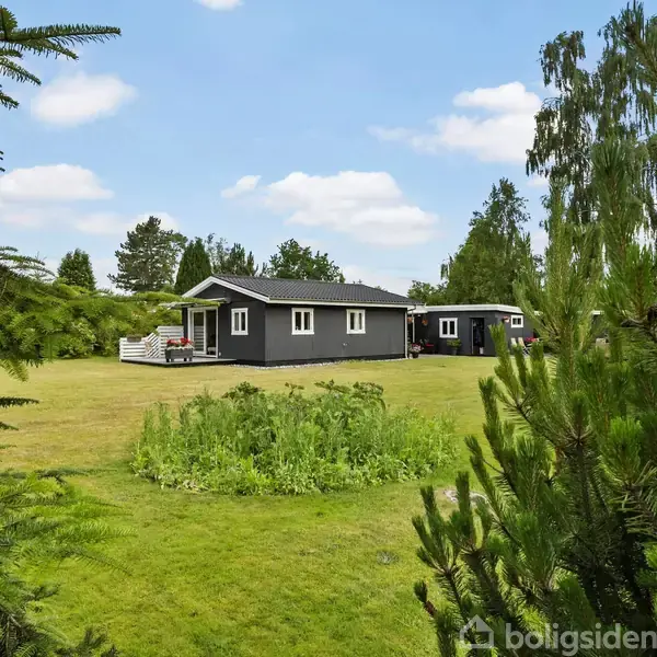 Et mørkegråt hus med hvide vinduer står på en stor grøn græsplæne omgivet af buske og træer under en blå himmel med skyer.