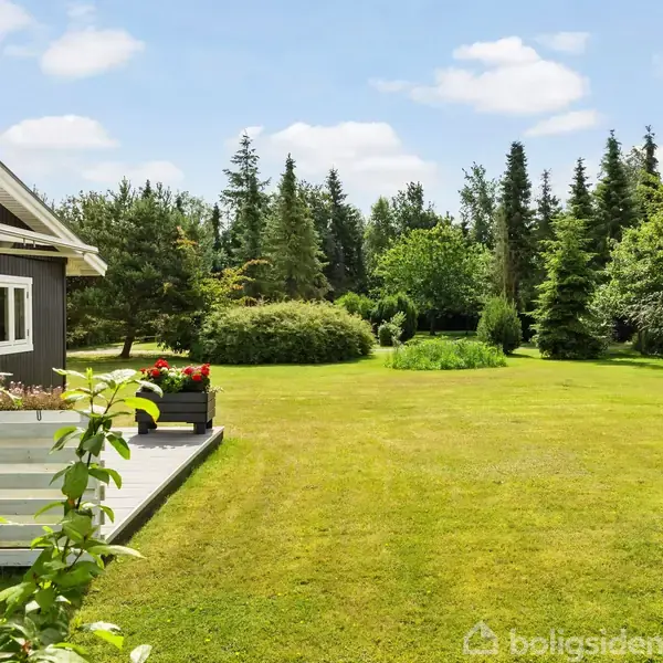 Hus med terrasse står stille i en stor, velplejet have omgivet af høje træer og buske under en lys, skyet himmel.