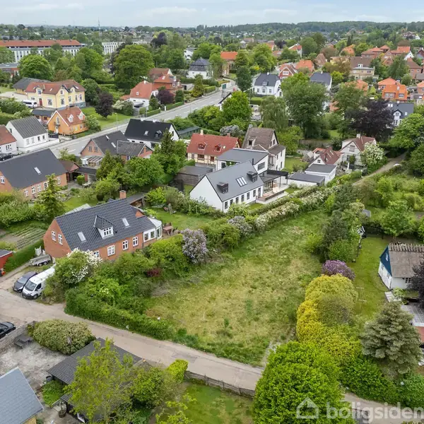 Bebyggelsen udgøres af huse med røde tage omgivet af frodigt grønt landskab. En åben græsmark dominerer midten, omgivet af veje og parkerede biler i et roligt villakvarter.