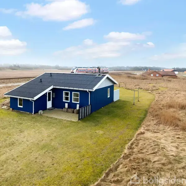 Et blåt hus med sort tag står på en grøn græsplæne omgivet af åbne marker. Der ses flere huse i det fjerne under en blå himmel med skyer.