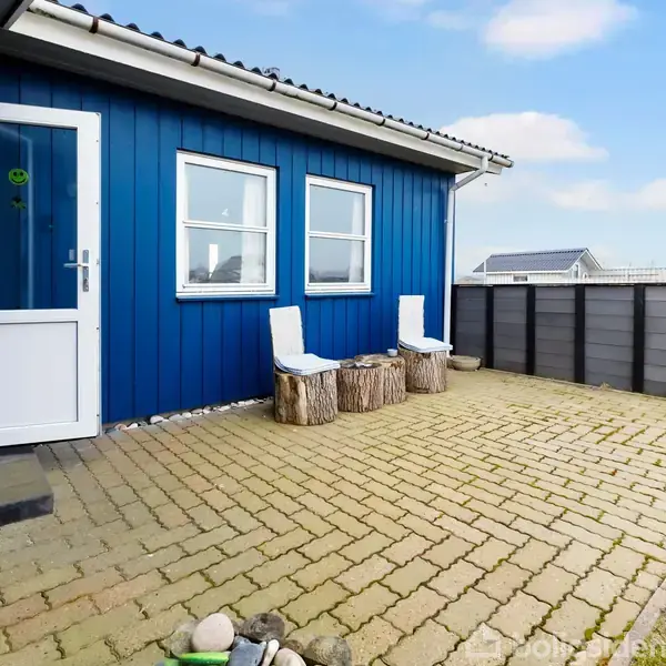 Blåt hus med to vinduer, hvide døre, og to hvide stole på træstubbe. Beliggende på en stenbelagt terrasse med græsplæne i baggrunden.