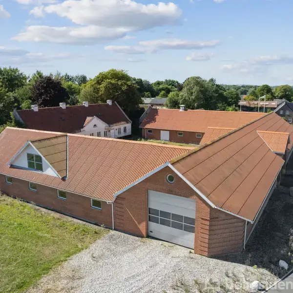 En stor bygning med rødt tegltag og garageport står ved en landevej. Omkring er der grønne træer og anden bebyggelse, alle under en himmel med spredte skyer.