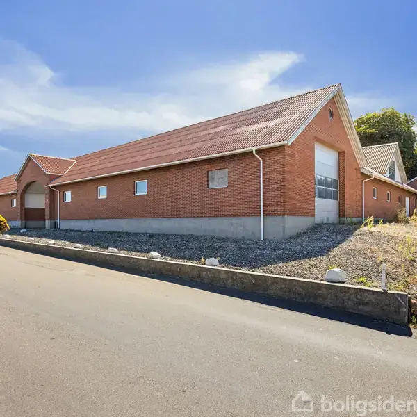 En stor, rød murstensbygning med skråt tag står stille ved en asfalteret vej. Den er omgivet af en grusbelagt gård og træer i baggrunden under en blå himmel.