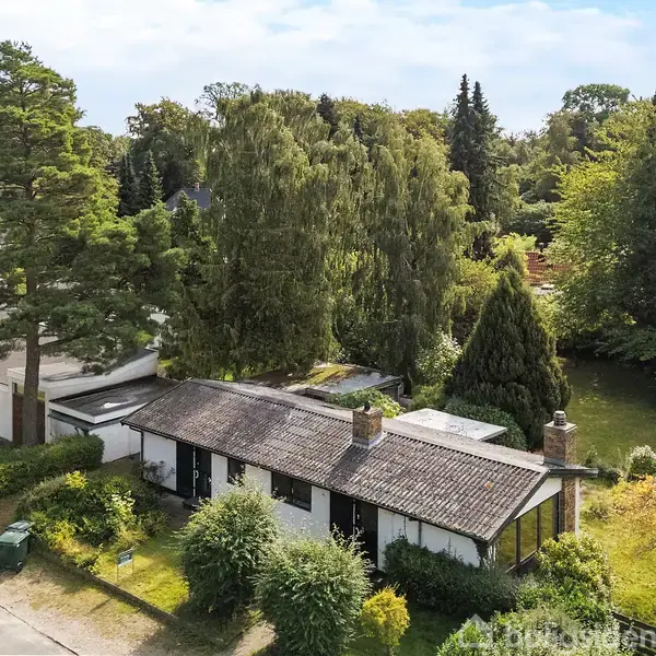 Et enfamilieshus med fladt tag står blandt frodige træer og buske. Huset er omgivet af en grøn have i et skovrigt område, hvor flere huse kan ses i baggrunden.