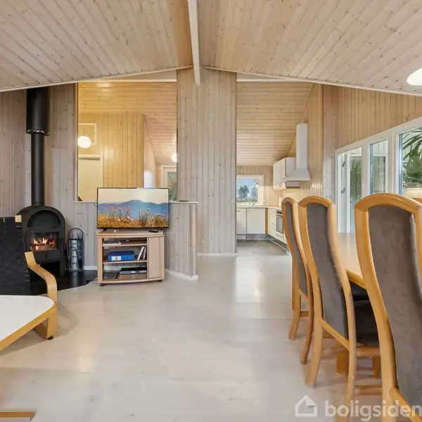 Spisestue med lang, træbehandlet bord og stole til højre, en sofa og sofabord til venstre, en brændeovn i hjørnet, fjernsyn på møbel og køkken i baggrunden.