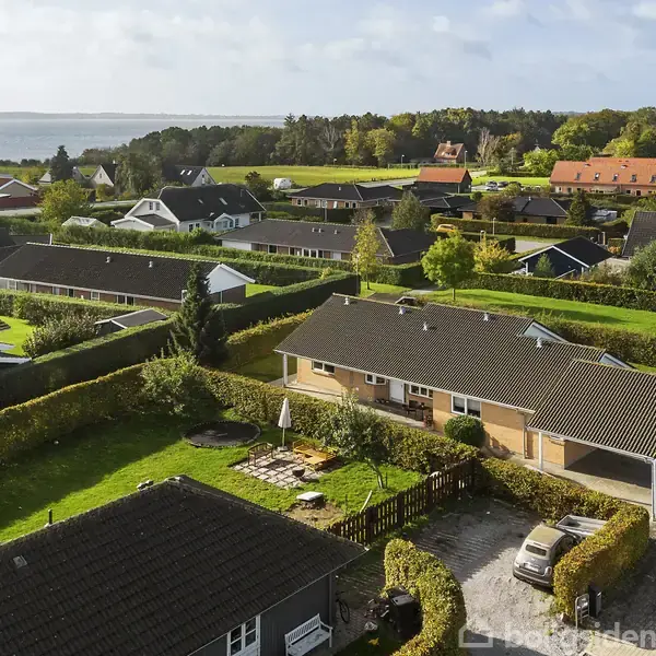Et parcelhus ses ovenfra med en velplejet have. Der er en terrasse med parasol og en bil i indkørslen. Omkringliggende huse og grønne marker ligger i baggrunden tæt ved kysten.