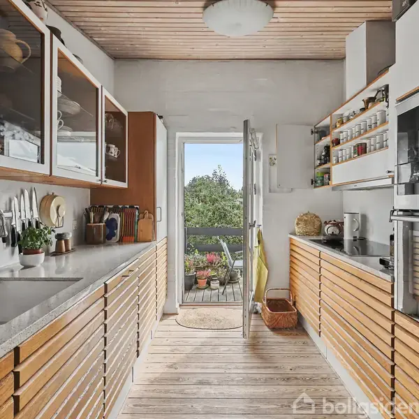 Et moderne køkken med træskuffer og åbne hylder fyldt med krydderier. Der er en vask til venstre og en indbygget ovn til højre. Dør åbner til balkon med planter.