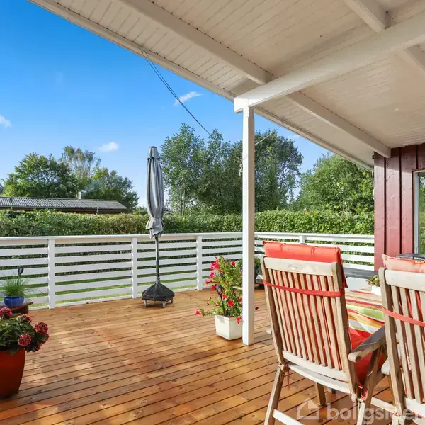 En træterrasse med spisebord og stole, omgivet af grønne buske og træer under en blå himmel. En parasol står ved siden af blomster, der giver farve til terrassen.