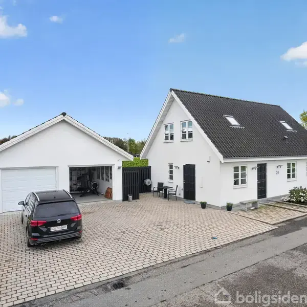 Et hvidt hus med sort tag og garage ligger i et villakvarter. En bil holder parkeret foran garagen, og der er skraldespande langs stien i forhaven.