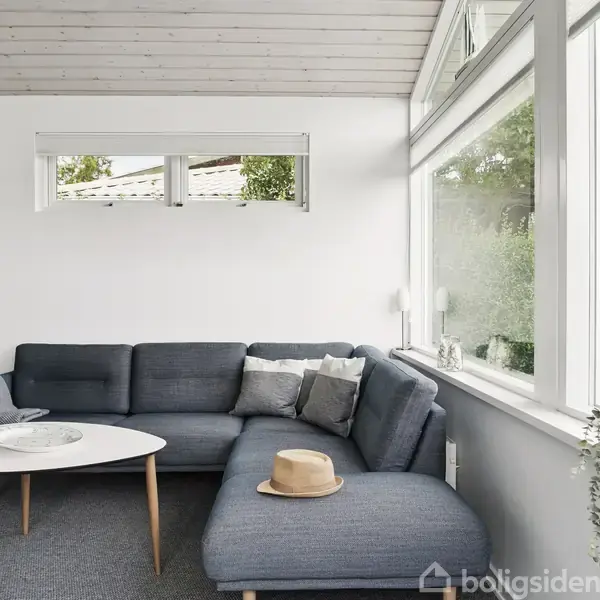 Sofa i blå nuancer står i stuehjørne med et rundt hvidt sofabord foran. Store vinduer giver udsigt til grønne buske. En hat ligger på sofaen.