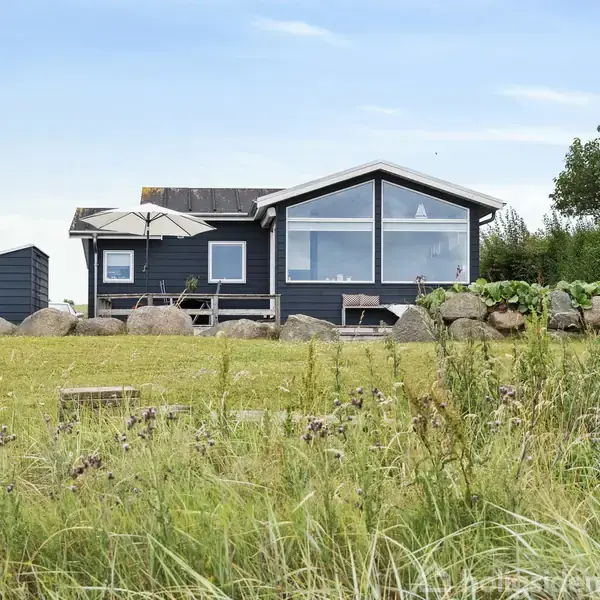 Et sort sommerhus med store vinduer står på en græsplæne omgivet af klippesten. En parasol dækker terrassen, mens træer og buske udgør den bagvedliggende have.