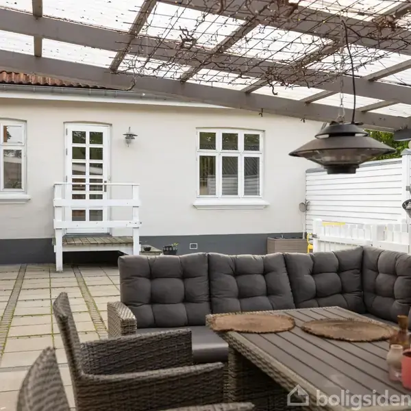 En grå polstret sofa står på en flisebelagt terrasse under en pergola. I forgrunden ses et bord og stole, omgivet af hvidmalede vægge med vinduer og en dør.