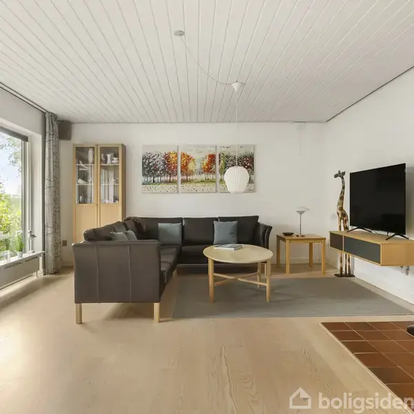En stue med en sort sofa og et rundt sofabord foran, har store vinduer til venstre med udsigt til en have. På væggen hænger et TV, og en brændeovn er til højre.