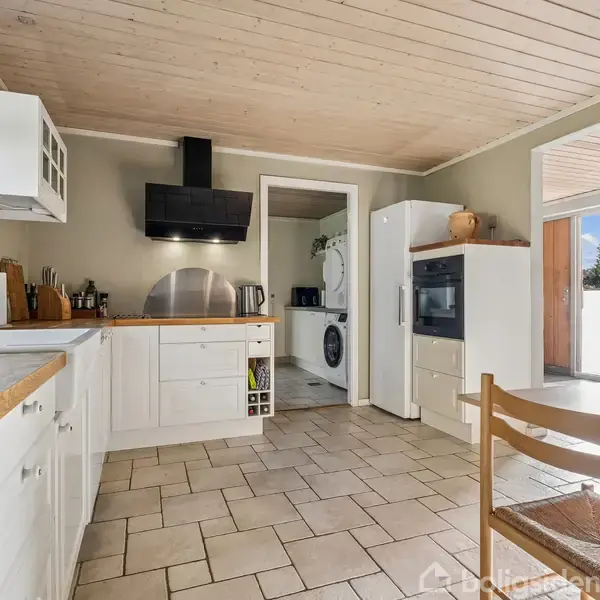 Køkken med hvide skabe og træbordplader, hvor en spiseplads står centralt. En dør åbner til et bryggers med vaskemaskine. Store vinduer bringer lys ind i rummet.
