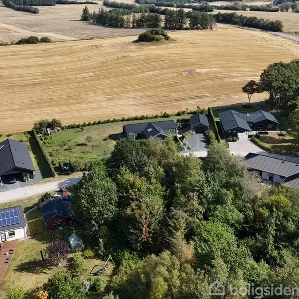 Huse med mørke tagsten ligger langs en vej, omgivet af frodig skov og marker. En bygning har solpaneler på taget. Marker strækker sig ud i det fjerne.