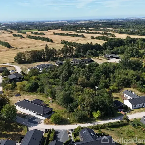 Et boligområde med huse omgivet af grønne træer og marker strækker sig til horisonten under en blå himmel på en solrig dag.