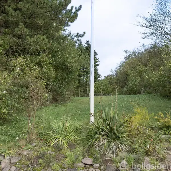 En hvid flagstang står i midten af en frodig have med grønne buske og træer rundt om, mens græsset dækker jorden.