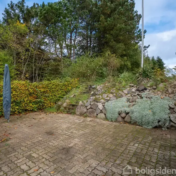 En lukket parasol står stille på en flisebelagt terrasse omkranset af grønne buske og træer. En stengrå mur med planter løber langs en skråning i baggrunden.