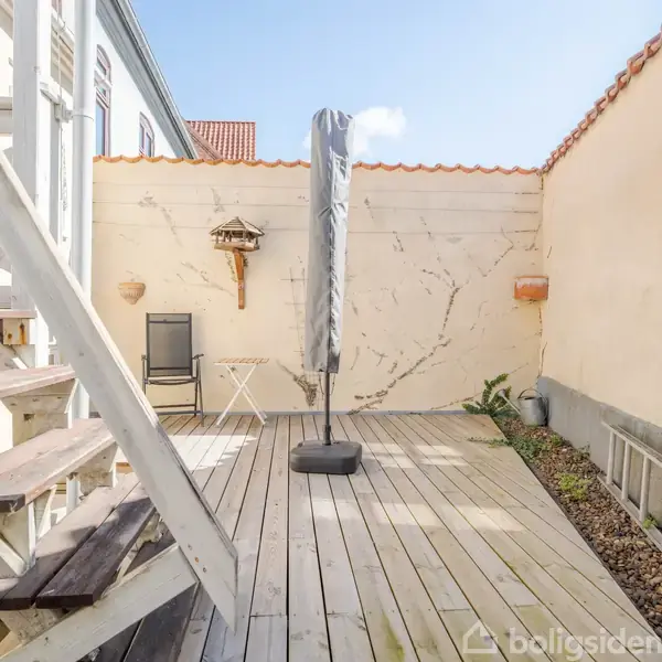 Terrasse med parasol i midten, omgivet af en træplatform. Til venstre en trappe, til højre planter. Væggen bagved har en stol, bord og fuglehus. Ingen tekst synlig.