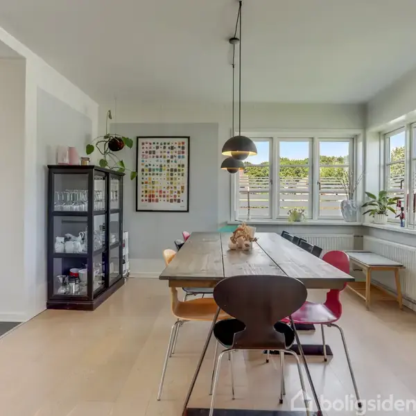 Et spisebord med stole står i et lyst rum med store vinduer og planter i vindueskarmen. Der hænger lamper over bordet, og der ses en trappe i baggrunden.