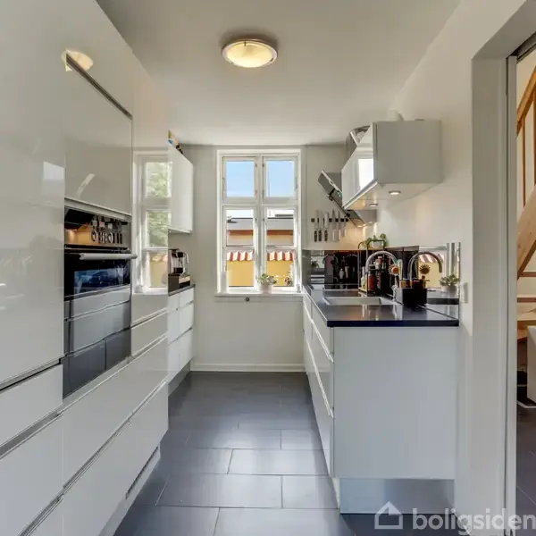 Et moderne køkken med hvide, glatte skabe og sorte bordplader. Køkkenet har en opvaskemaskine og komfur, vindue i baggrunden, og er ved siden af en trappe.