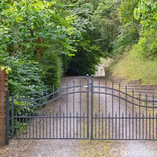 En sort metalport står lukket, flankeret af røde murstenssøjler. Bag porten løber en grusvej, omgivet af frodig grøn vegetation på begge sider. Nummeret 22 ses på venstre søjle.