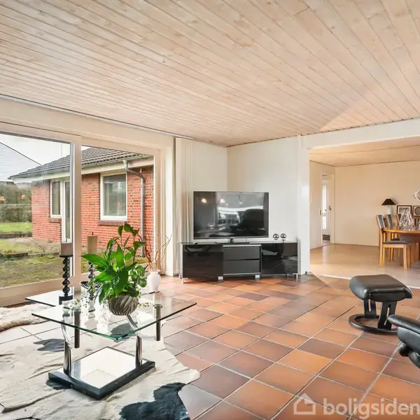 Stue med glasbord og grøn plante, lænestol ved glasdør, fladskærms-TV på sort reol, åben overgang til spisestue med spisebord, omgivet af murstenshus og have udenfor.