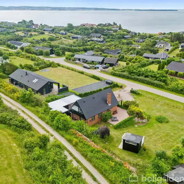 Hus med sort tag og skorsten beliggende i et grønt landskab. Omkringliggende vej fører til flere huse. I baggrunden kan man se havet.
