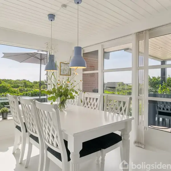 Et hvidt spisebord med seks stole er centralt placeret i et lyst rum. Bordet er pyntet med en vase med blomster. Store vinduer åbner op til en terrasse med udsigt over grønne omgivelser.