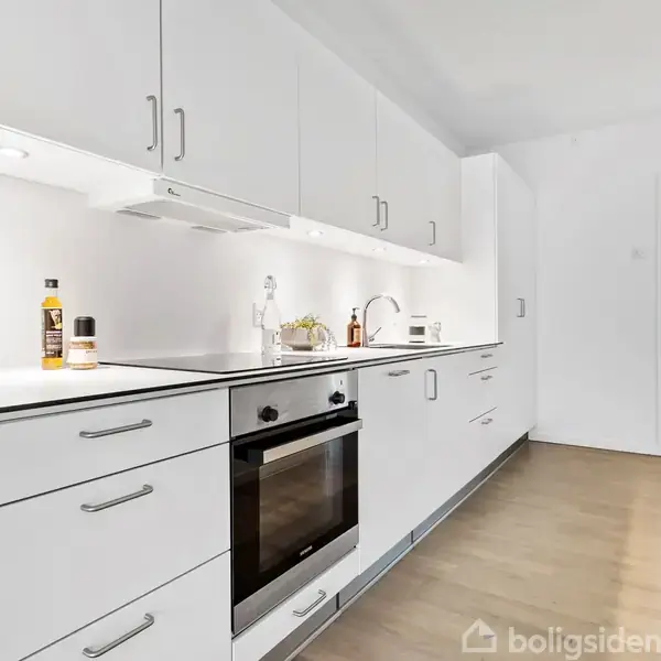 Hvidt køkken med moderne skabslåger og en indbygget ovn i aktion. Køkkenet er oplyst og ryddeligt, med en flaske og dekorative genstande på bordpladen, der fører til en stue.