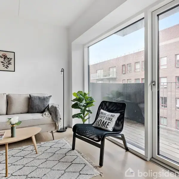 En moderne stue indeholder en grå sofa ved siden af et lille, rundt træbord. En lænestol med en pude påskrevet "THERE IS NO PLACE LIKE HOME" står ved store glasdøre, der leder til en altan med udsigt til en moderne bygning. Gulvtæppet er mønstret, og stuen er dekoreret med enkelte grønne planter.