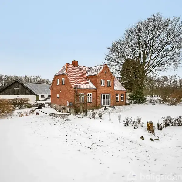 En rødstensvilla med skråt tag står stille i et snedækket landskab. Villaen er omgivet af buske og træer, med en flagstang tæt ved.