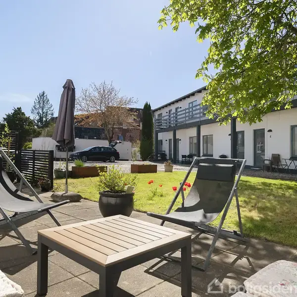 To liggestole står på en terrasse med et bord imellem dem. I baggrunden ses en græsplæne med et træ, og et moderne, hvidt hus med altaner.