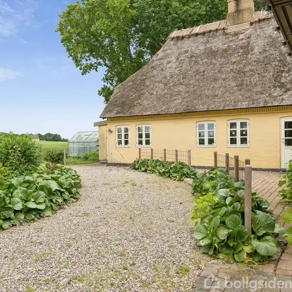 Gult, stråtækt hus langs en grussti omkranset af grønne planter. Baggrunden har en græsplæne med et drivhus og grønne marker under en blå himmel med træer rundt om.