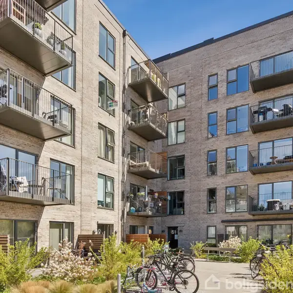 En moderne lejlighedsbygning med flere etager, altaner med planter, cykler parkeret foran og grøn beplantning omkring. Bygningen har store vinduer og er lavet af mursten.